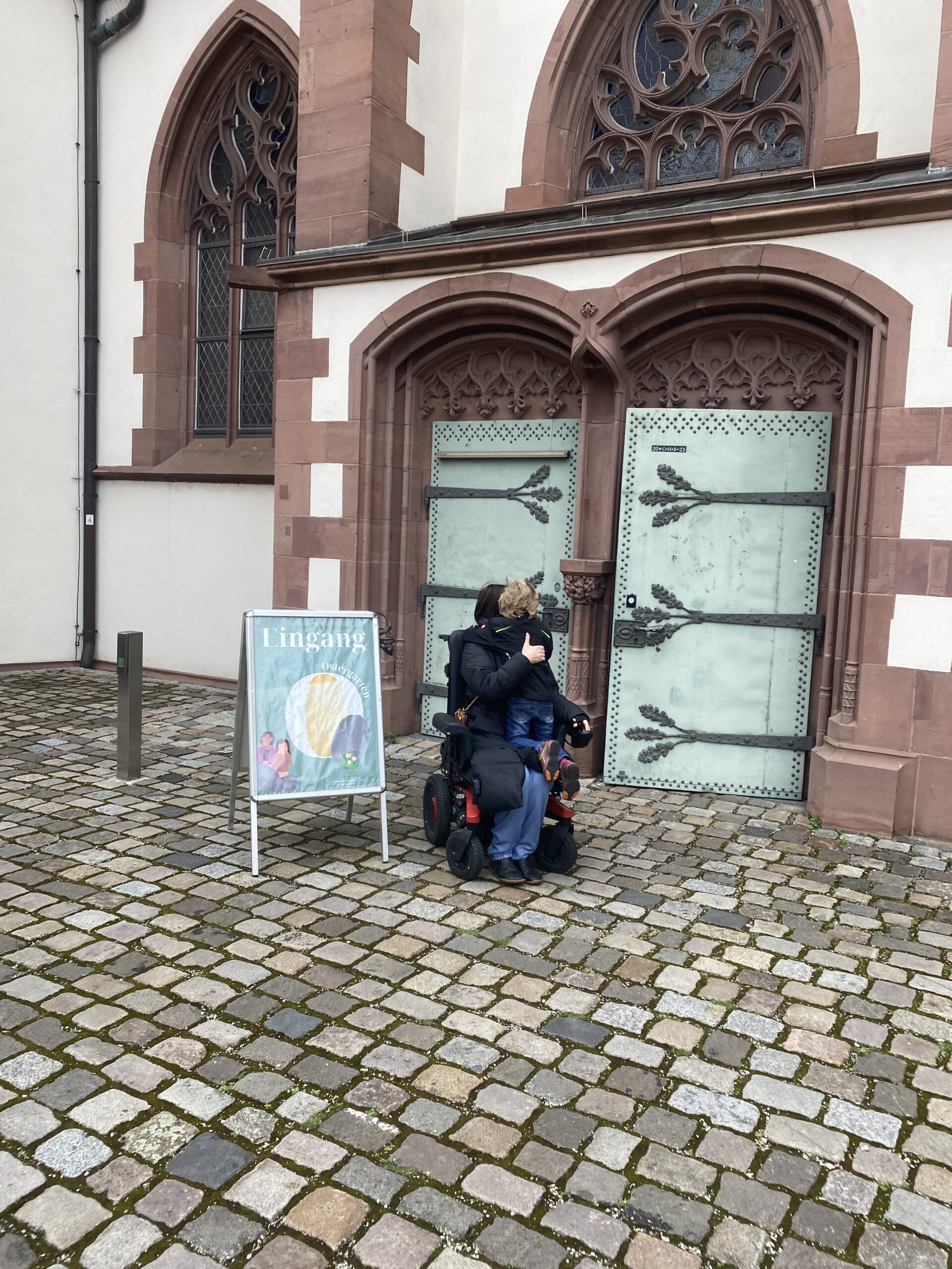 Ich stehe neben dem Schild: Eingang zum Ostergarten und daneben ist die Tür zur Jugendkirche. DAs Januarwunder kniet auf meinen Oberschenkeln 