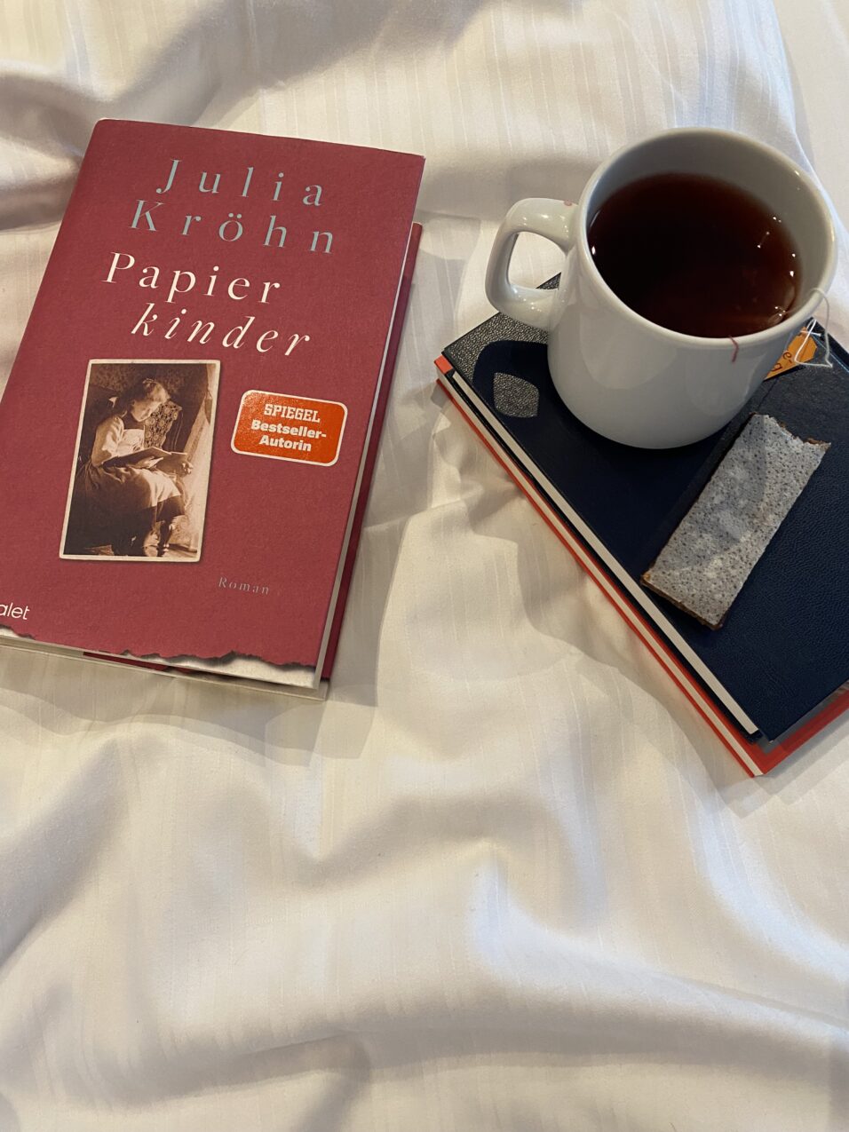 Auf dem Bett liegt links das Buch und neben steht auf zwei Büchern ein Tee und ein Fruchtriegel liegt dabei