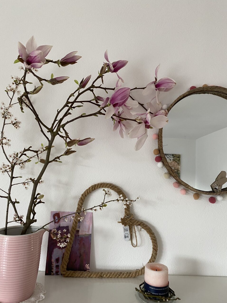 Aufgeblühte Magnoloen in einer Vase, vor der Wand. daneben steht noch weitere Deko und ein runter Spiegel hängt an der Wand